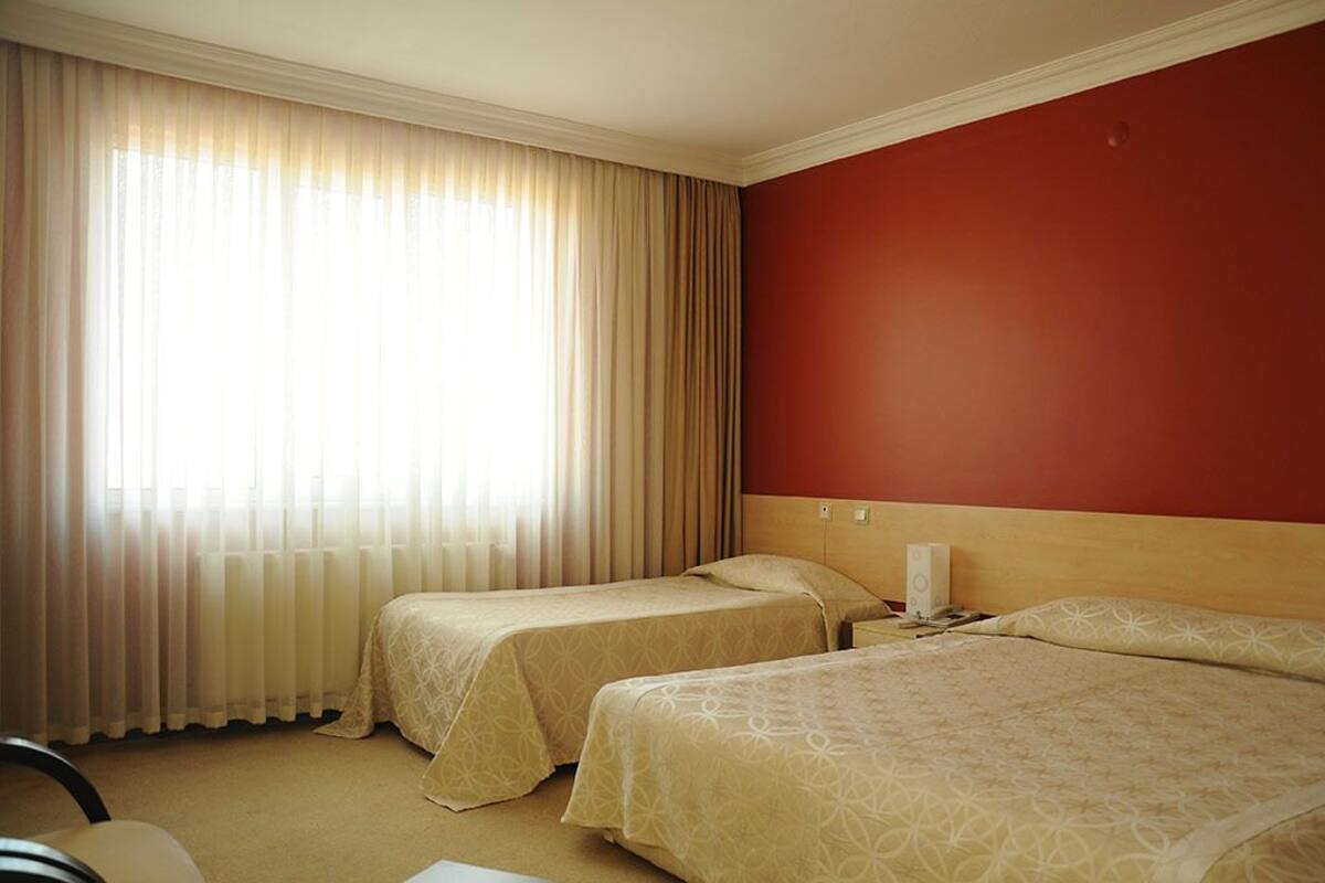 A hotel room with two beds, beige bedding, a red accent wall, and a large window with sheer curtains.