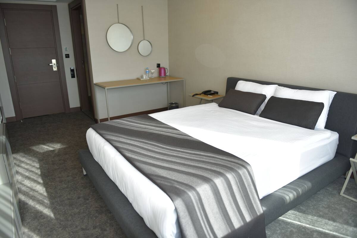 A hotel room with a large bed, a bedside table, a desk, a chair, a mirror, and a door.