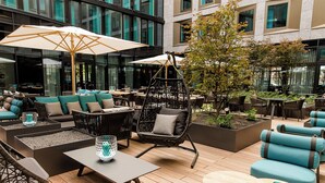 Un espace extérieur aménagé avec des chaises confortables, des tables et des parasols, entouré de verdure et bordé d’une architecture moderne.
