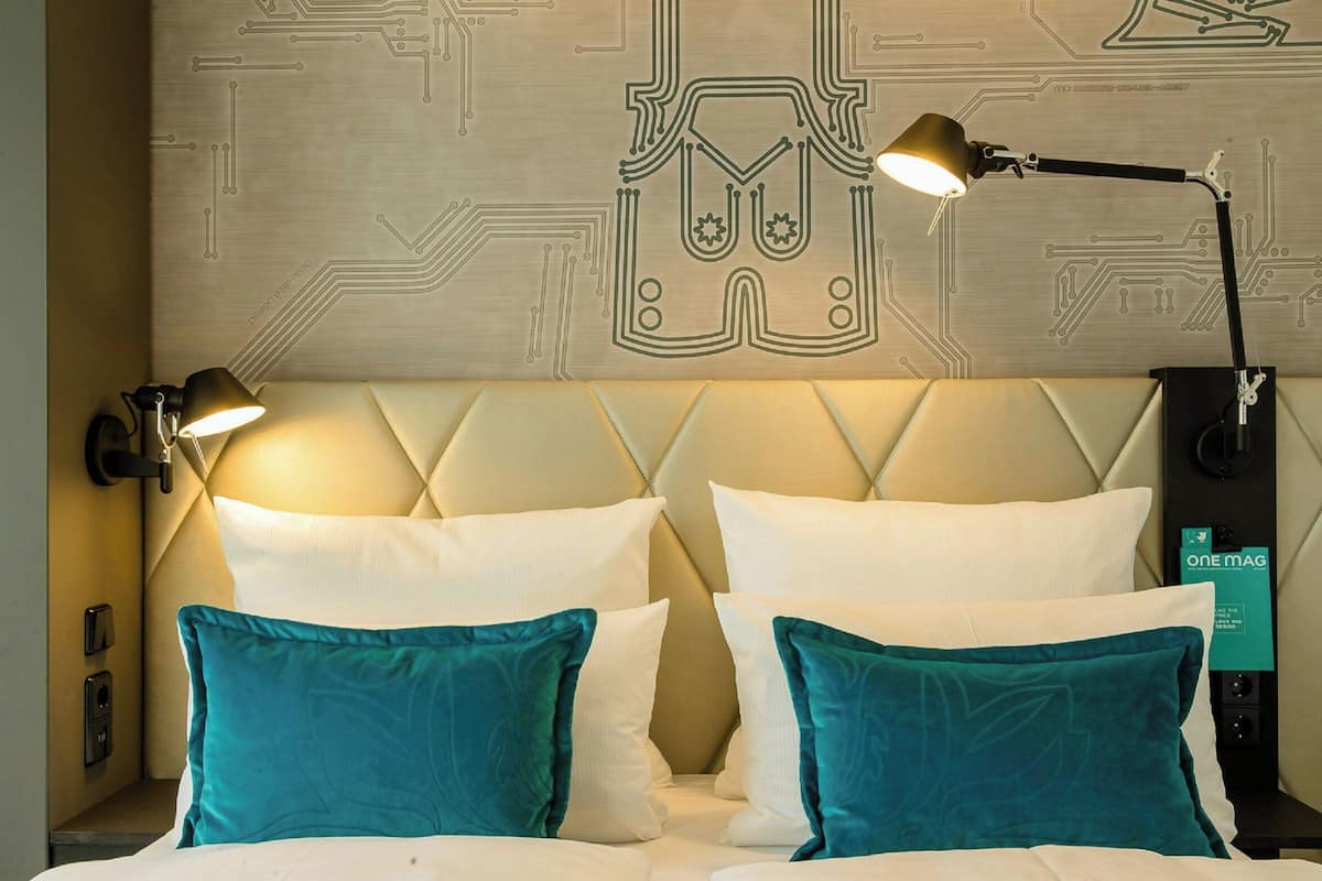Habitación de hotel con una cama, dos lámparas de noche y una pared con un diseño tipo placa de circuito.