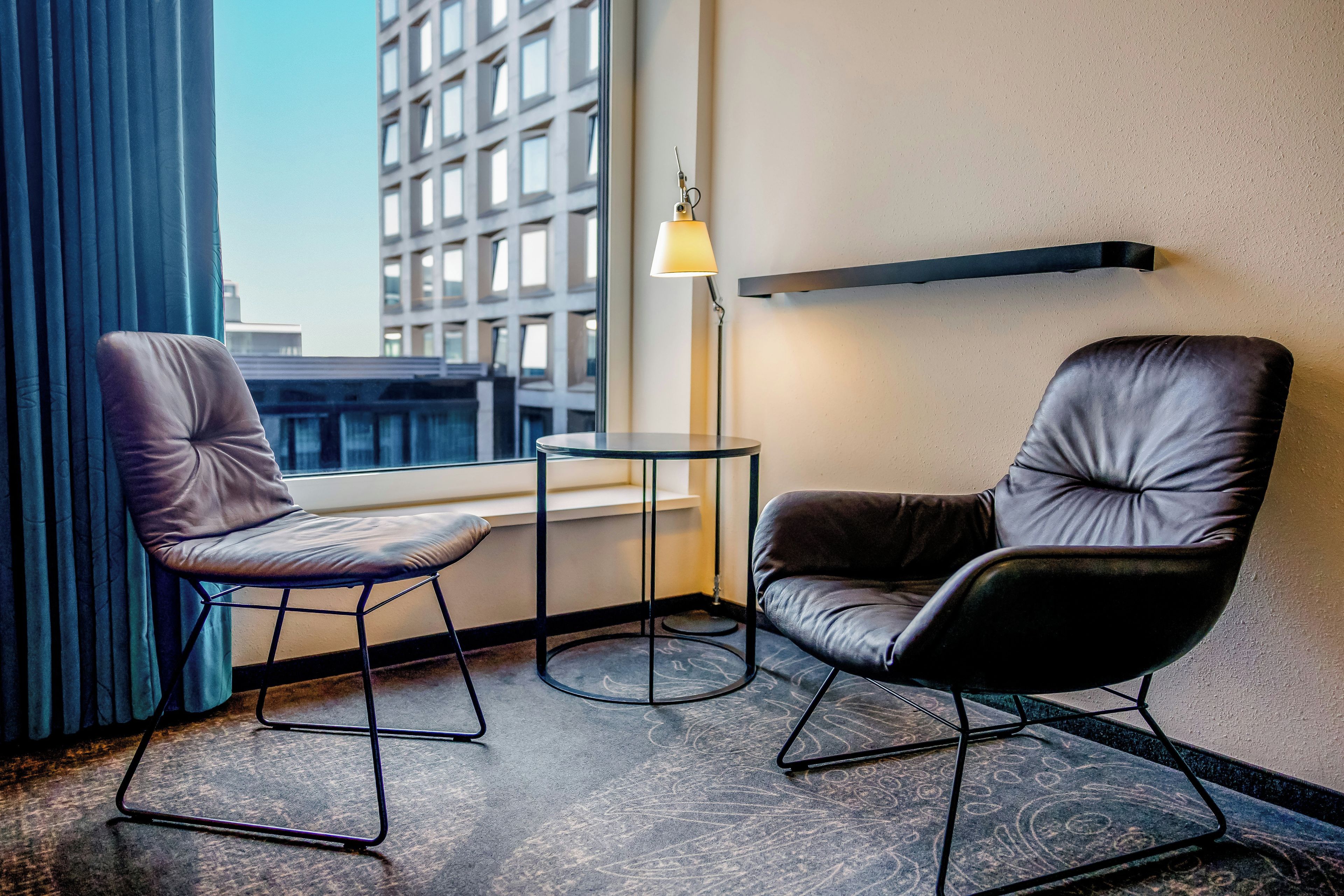 Habitación de hotel con dos sillones de cuero, una mesita, una lámpara de pie y vistas a un edificio a través de una ventana.