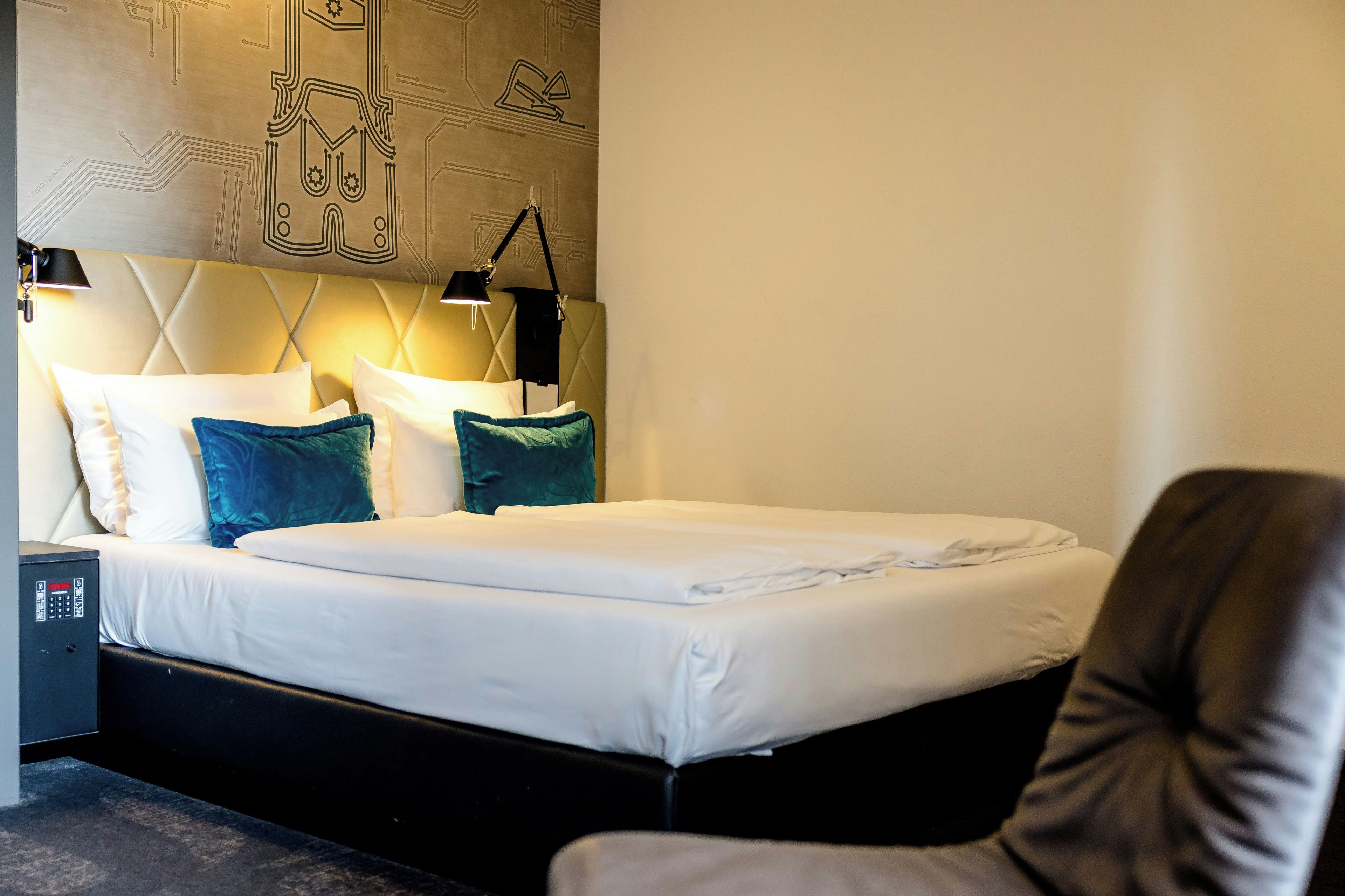 A hotel room with a bed, pillows, a desk with a lamp, and a wall with a circuit-like design.