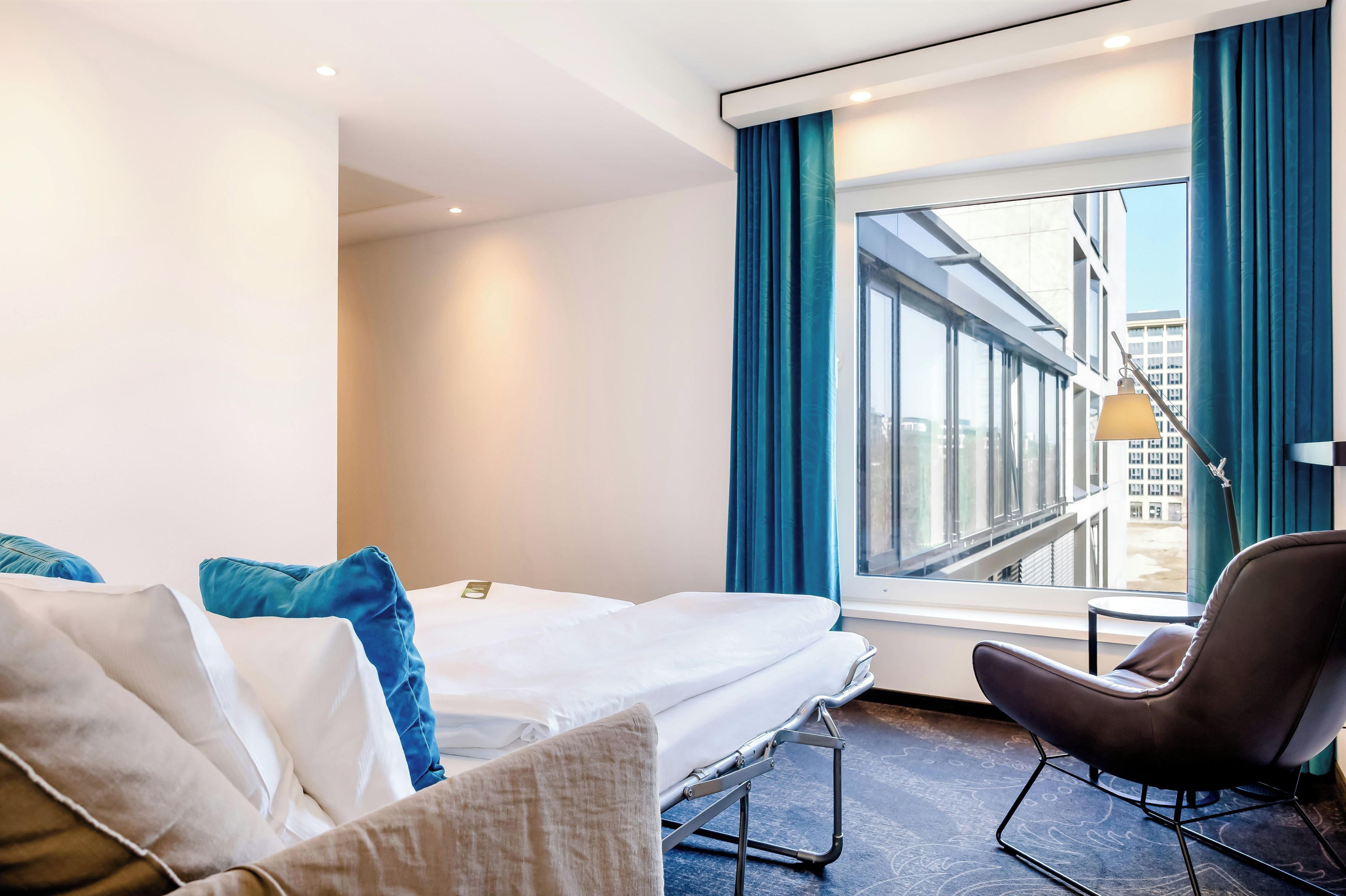 Hotellihuone, jossa on sänky, tuoli, ikkuna, josta on näkymä rakennuksiin, ja lamppu.