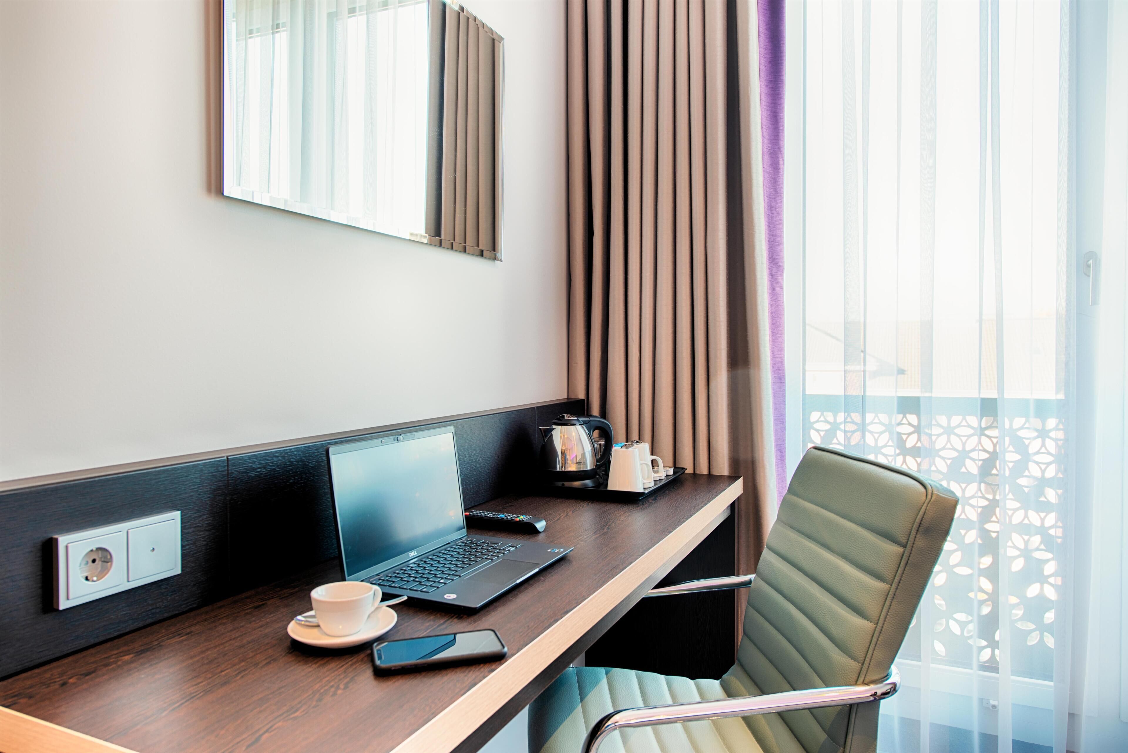 A hotel room with a wooden desk, a laptop, a smartphone, a cup, and a coffee maker.