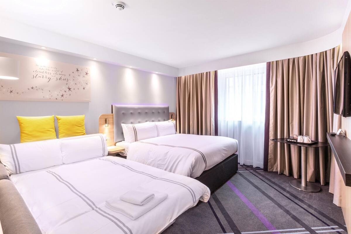 Une chambre d’hôtel avec deux lits, un téléviseur à écran plat, une table de chevet et une grande fenêtre avec des rideaux.