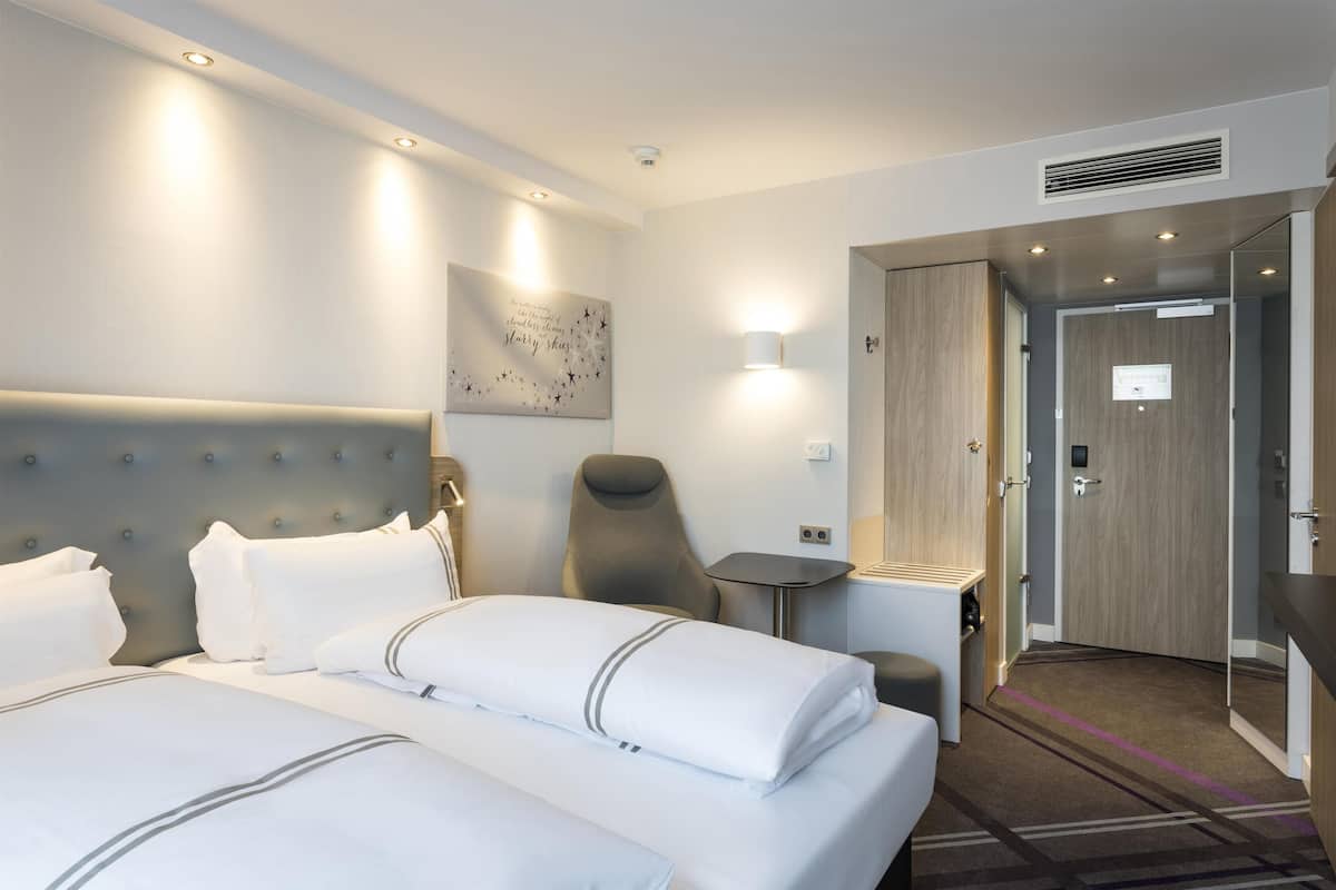 Une chambre d’hôtel avec un grand lit, une chaise, une petite table, une lampe fixée au mur et une porte donnant sur une autre pièce.