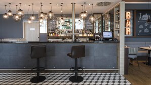 Una zona de bar con suelo a cuadros, dos taburetes de bar, lámparas colgantes y una barra con un ordenador y estanterías llenas de botellas.