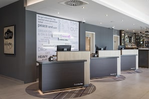 A hotel reception area with a modern design, featuring a large wall with various words and phrases, a reception desk, and a bar area. - Premier Inn Freiburg City Süd (Freiburg im Breisgau)