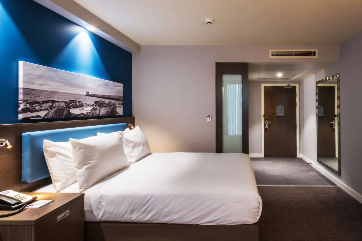 A hotel room with a bed, a nightstand, a phone, a wall-mounted picture, and a door leading to another room.