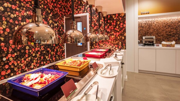 Un'area buffet con diverse opzioni alimentari, tra cui un contenitore blu con l'etichetta 'bacon' e un contenitore rosso con l'etichetta 'bake'.