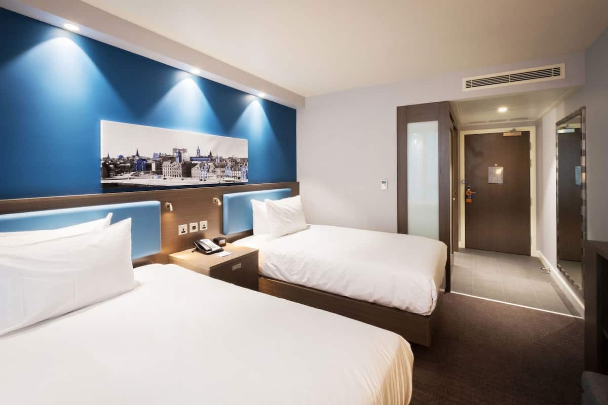 A hotel room with two beds, a nightstand with a phone, a wall-mounted picture, and a door leading to another room.