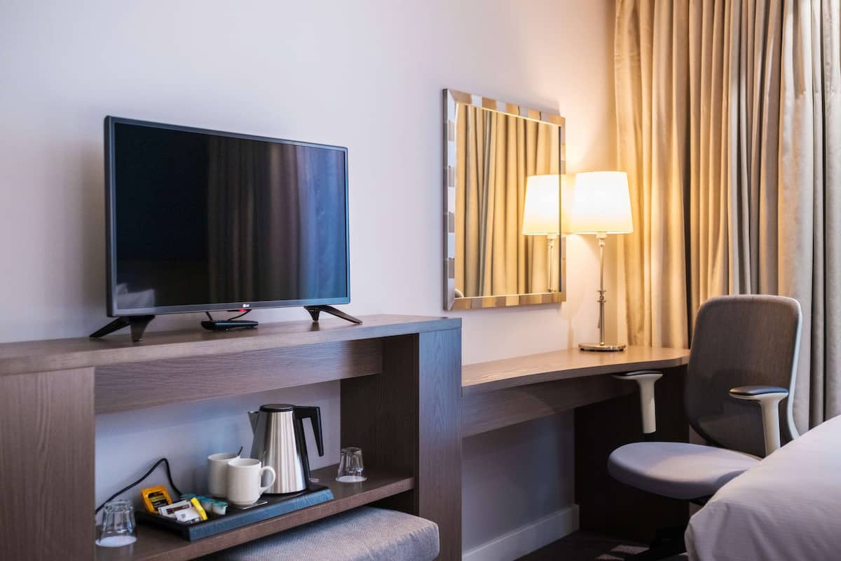 A hotel room with a flat-screen TV on a wooden stand, a desk with a lamp, a chair, and a bed.