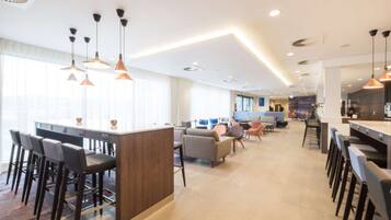 A modern hotel lobby with a central bar area, seating arrangements, and a visible kitchen in the background.