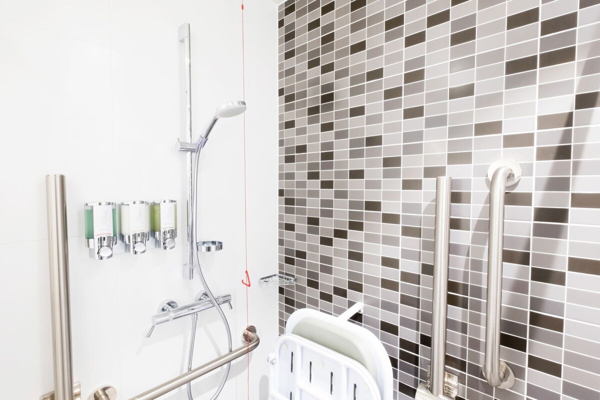 A bathroom with a shower, handrails, a white bathtub, and a tiled wall.