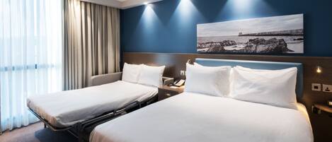Habitación de hotel con dos camas, un ventanal con cortinas, un cuadro de una escena costera y una mesita de noche con teléfono.