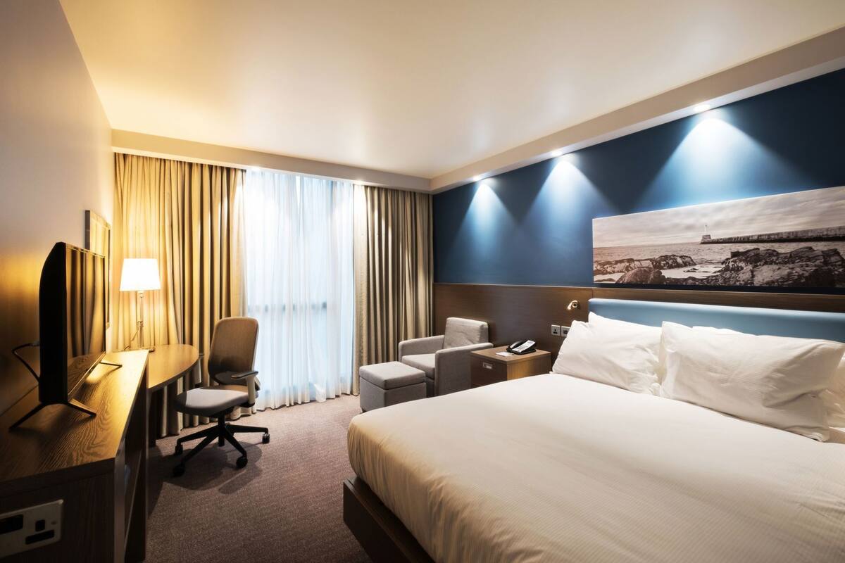 A hotel room with a bed, desk, chair, and a painting on the wall.