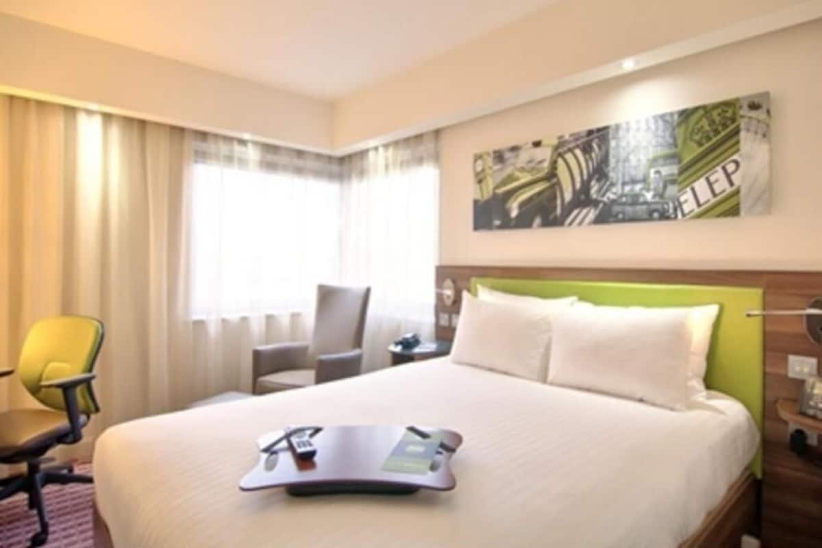 A hotel room with a bed, a desk, a chair, a window with curtains, and a wall-mounted artwork.