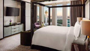 A modern hotel room with a large bed, a TV, a desk with a vase, and a view of a cityscape.