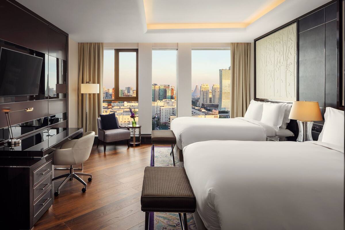 Ein Hotelzimmer mit zwei Betten, einem Schreibtisch, einem Stuhl, einem Fernseher und Blick auf die Stadt.