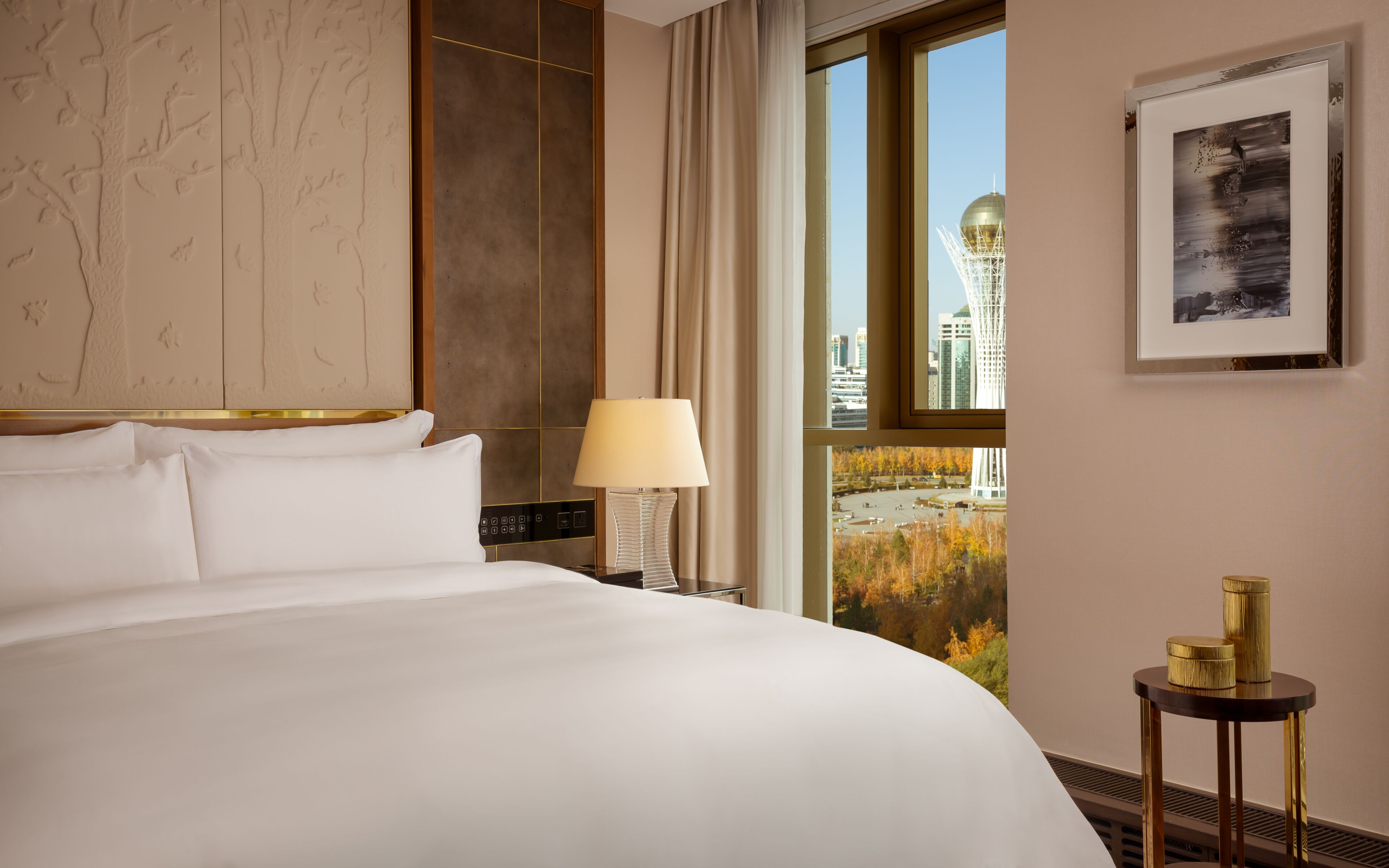 Habitación de hotel con una cama grande, una mesita de noche con lámpara, una pequeña mesa con objetos decorativos, un cuadro enmarcado y una ventana con vista a la ciudad.