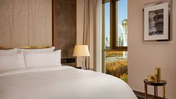 Habitación de hotel con una cama grande, una mesita de noche con lámpara, una pequeña mesa con objetos decorativos, un cuadro enmarcado y una ventana con vista a la ciudad.