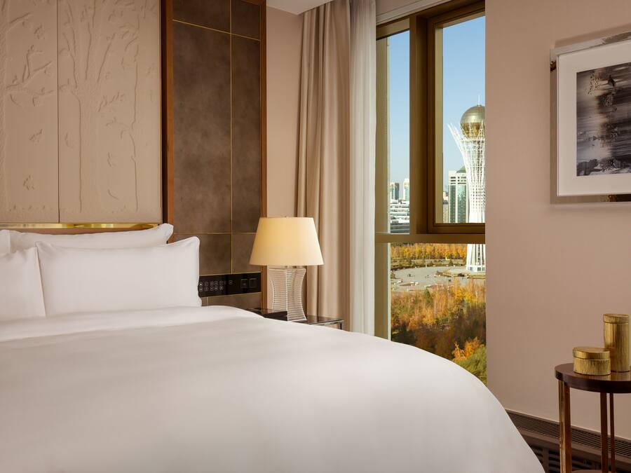 Habitación de hotel con una cama grande, una mesita de noche con lámpara, una pequeña mesa con objetos decorativos, un cuadro enmarcado y una ventana con vista a la ciudad.