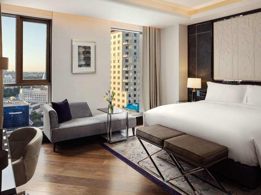 A modern hotel room with a large bed, a sofa, a small table with a vase, and a view of a cityscape through large windows.