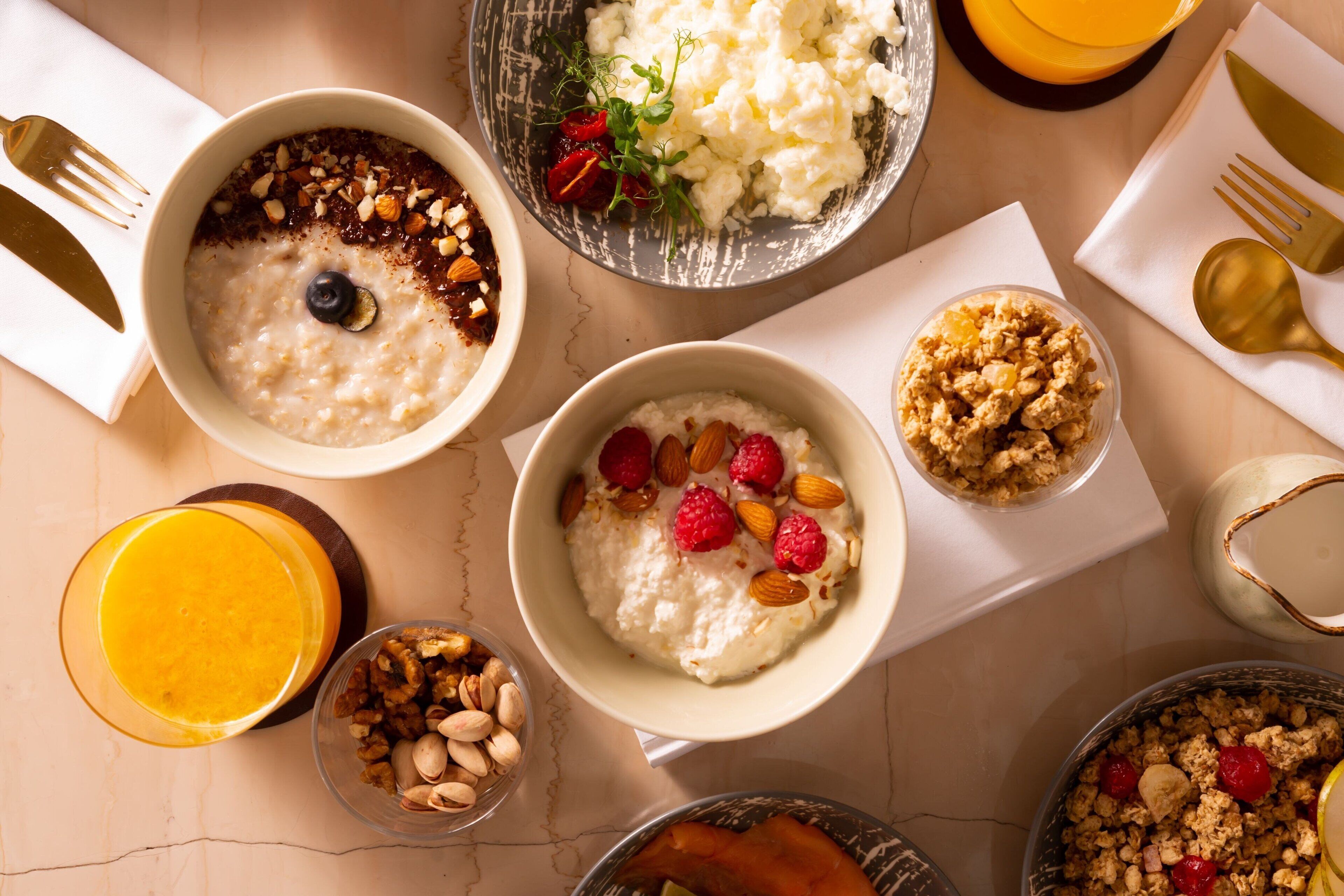 Variedad de platos para el desayuno, como avena, yogur y granola, sobre una mesa con cubiertos y un vaso de jugo de naranja.