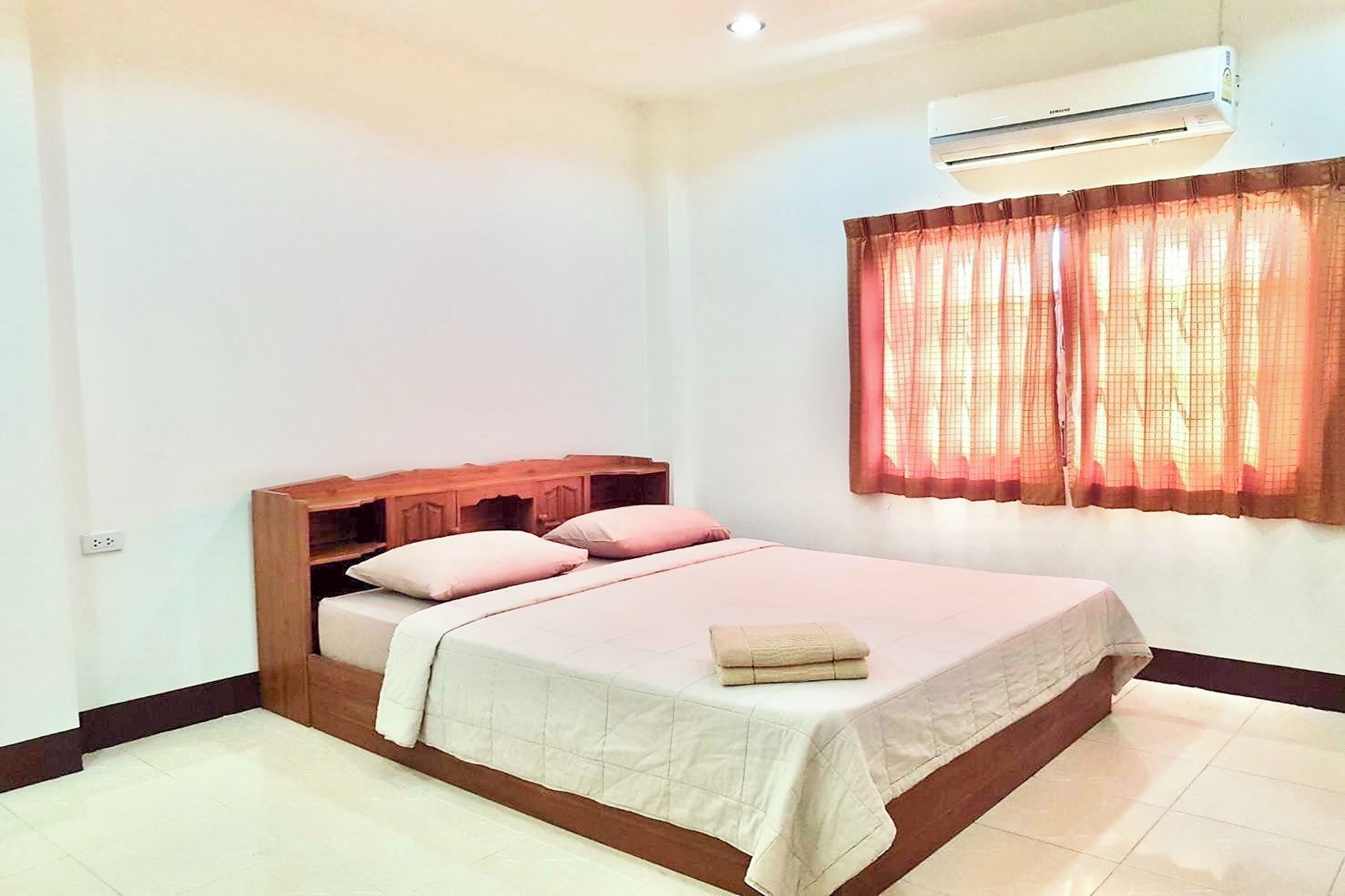 Une chambre à coucher bien rangée, avec une tête de lit en bois, un lit recouvert d’une couverture beige, des coussins roses et une serviette pliée posée sur le lit.