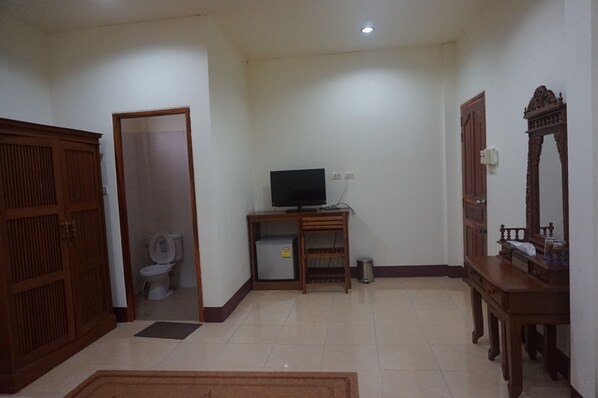 A room with a wooden wardrobe, a television on a stand, a small refrigerator, and a mirror on a wooden cabinet.