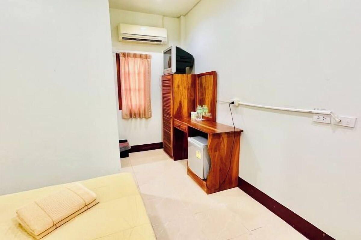 A small, clean room with a bed, a wooden desk, a small refrigerator, and an air conditioning unit.