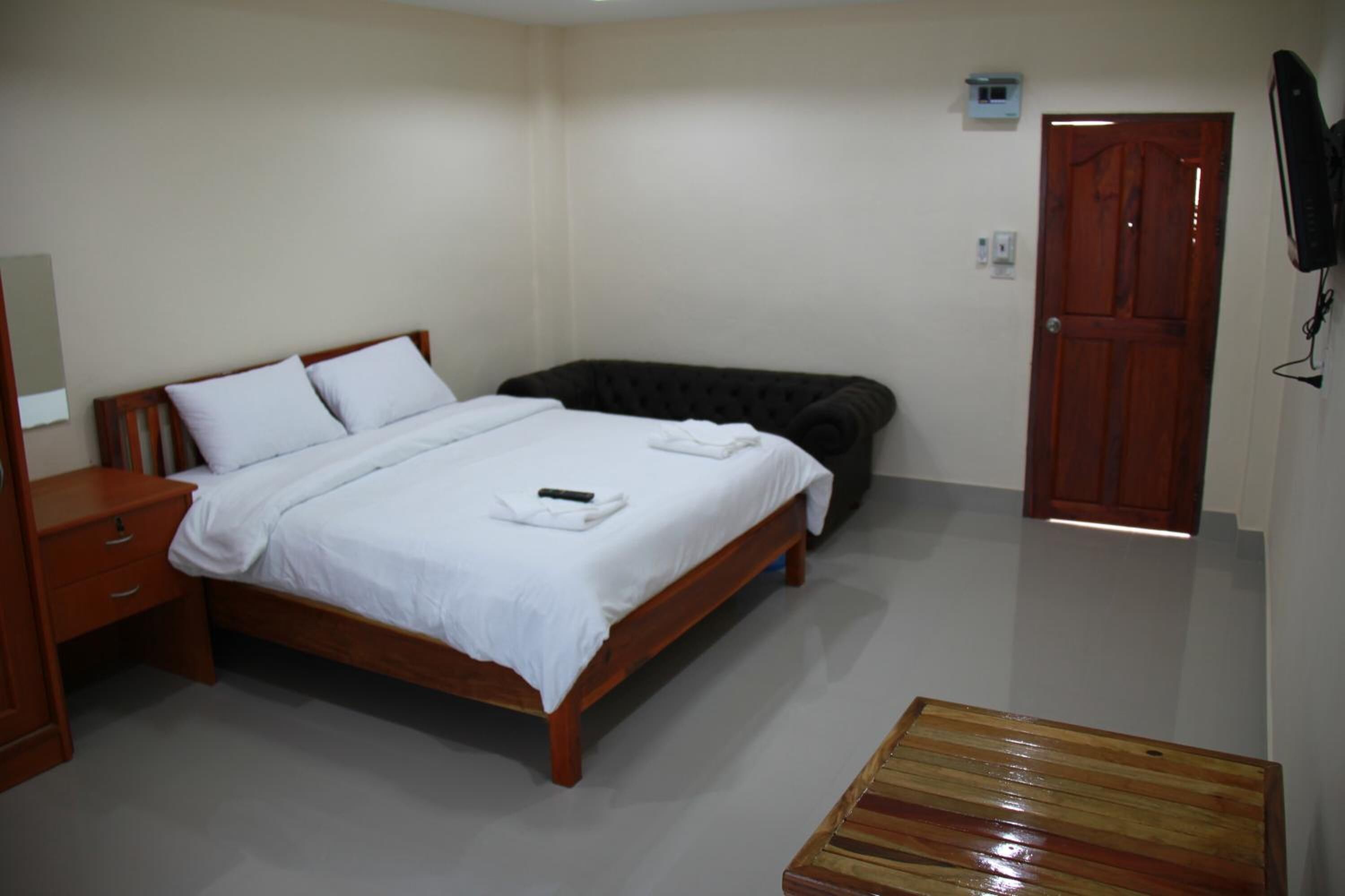 Un dormitorio con una cama de madera, una mesita de noche, un banco de madera, una puerta y un televisor instalado en la pared.