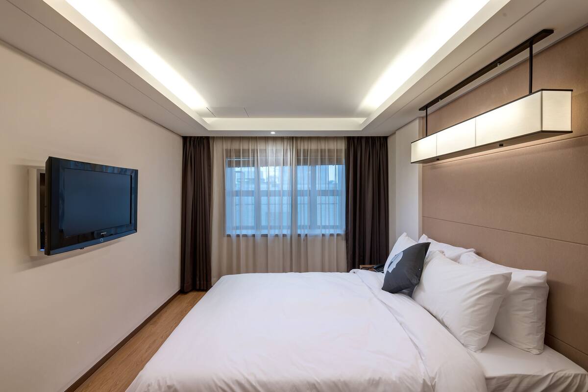 A modern hotel room with a large bed, a flat-screen TV mounted on the wall, and a window with curtains.