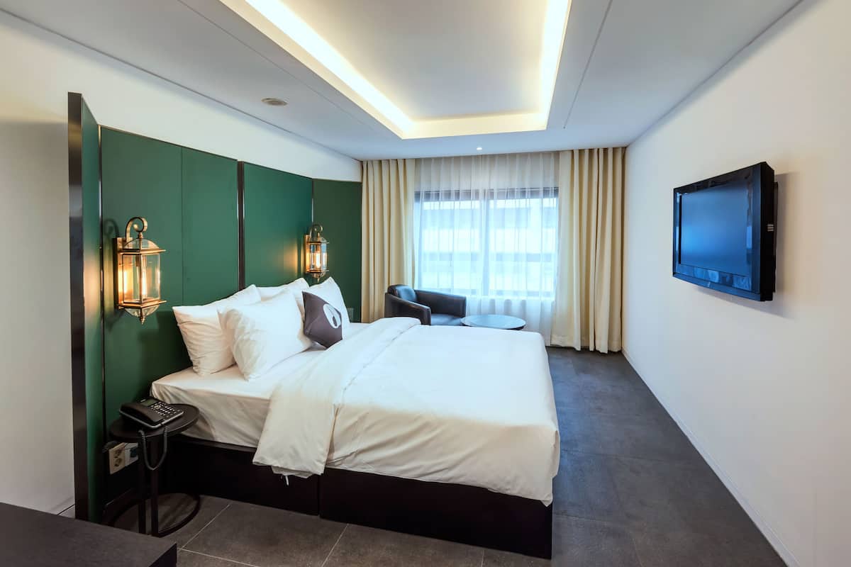 A hotel room with a large bed, two wall-mounted lamps, a flat-screen TV, a small table with a phone, and a window with curtains.