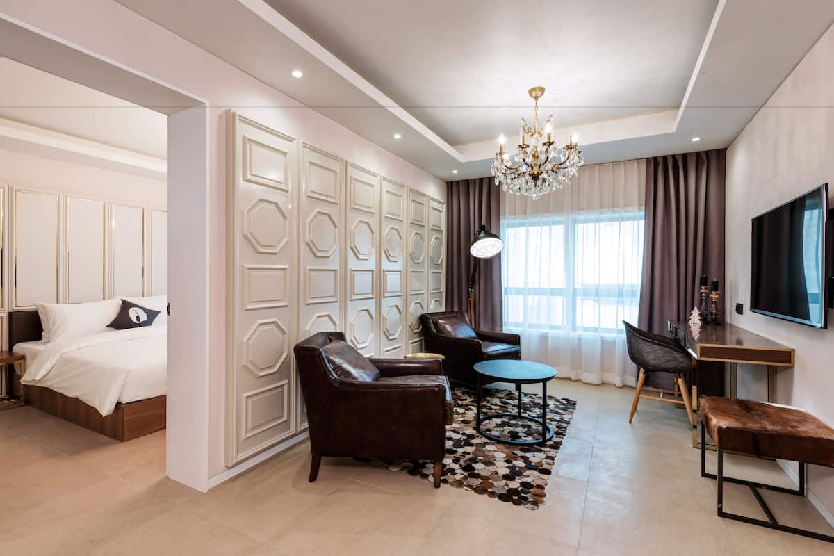 A modern hotel room with a large bed, a seating area with a sofa and armchairs, a small round table, and a TV mounted on the wall.