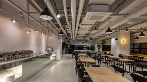 A modern cafeteria with long tables and chairs, a buffet area with food containers, and pendant lighting. - Hotel The Designers Yeouido (Seoul)