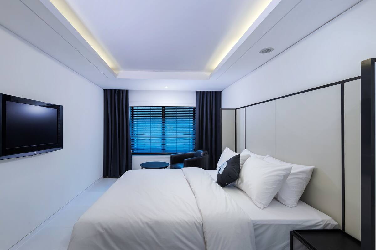 A modern hotel room with a large bed, a TV mounted on the wall, a small round table, and a window with blinds.