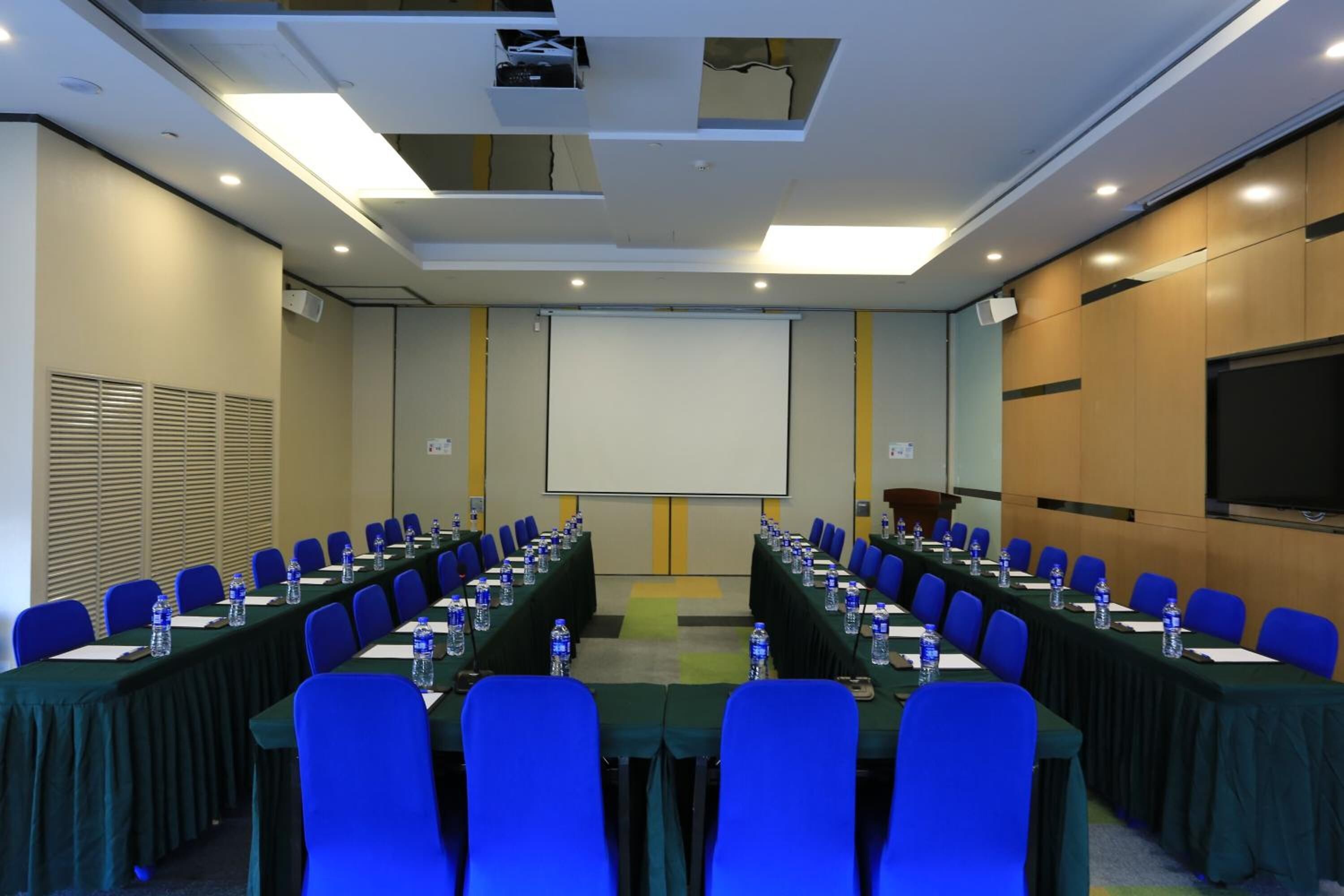 Een vergaderzaal met een lange tafel, blauwe stoelen, waterflesjes en een projectiescherm.