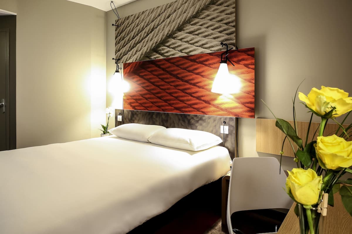 A hotel room with a bed, a nightstand, a wall-mounted lamp, and a vase with yellow flowers.