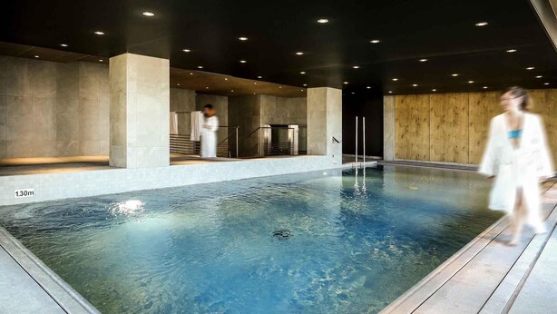 Une grande piscine intérieure à l’eau claire, entourée d’une terrasse en bois et de murs en béton.