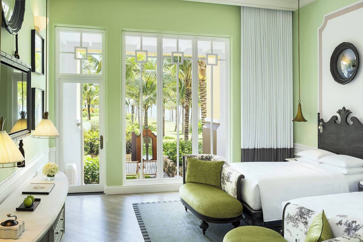 Ein Hotelzimmer mit einer großen Glasschiebetür, die auf einen Balkon mit Palmen führt. Es gibt zwei Betten mit grün-weißer Bettwäsche, einen Schreibtisch mit einer Lampe und einen Spiegel an der Wand.
