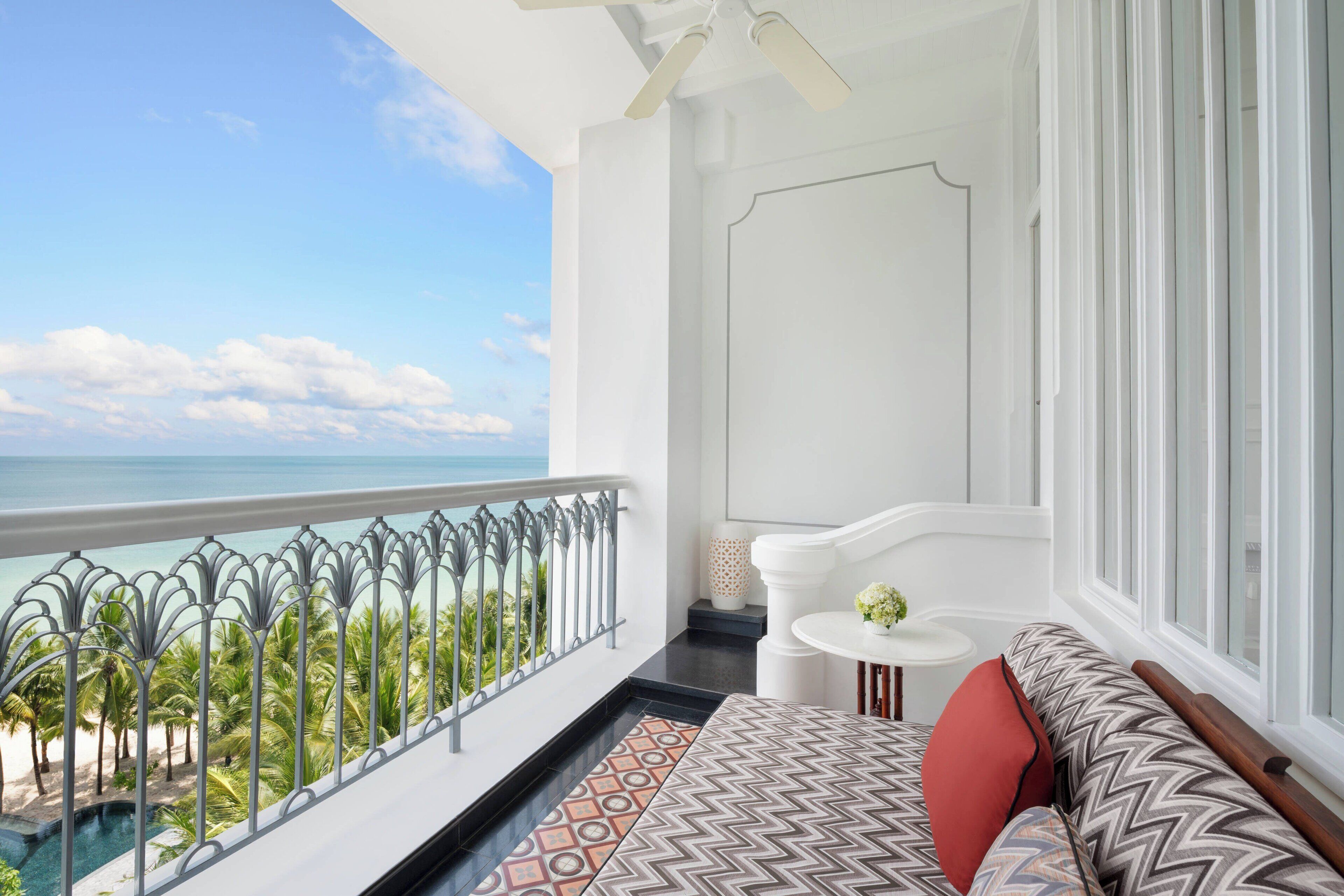 A balcony with a view of the ocean and palm trees, a white sofa with patterned cushions, and a small round table with a vase.