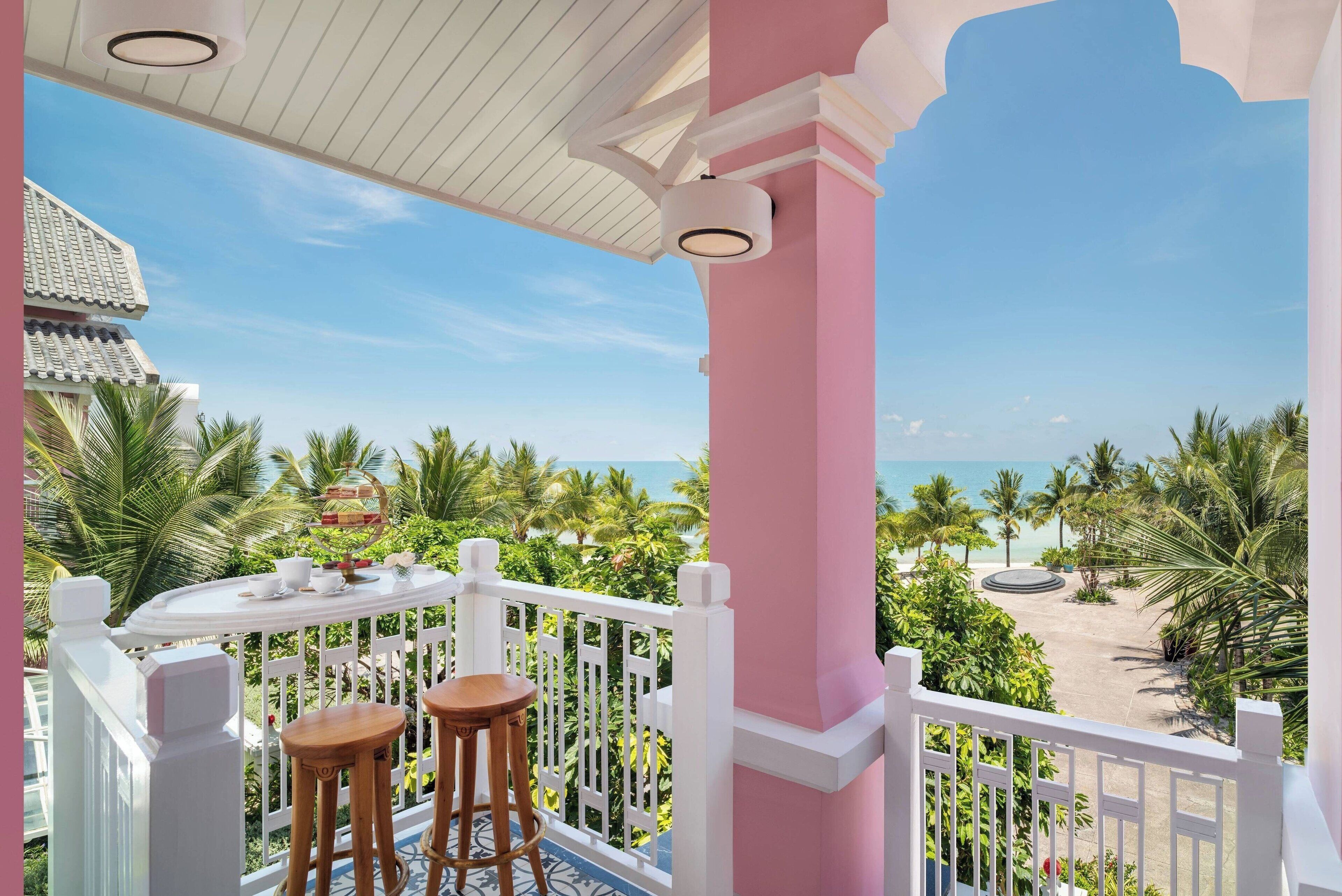 Un balcón con vista a la playa, barandillas blancas y una mesa con dos taburetes.