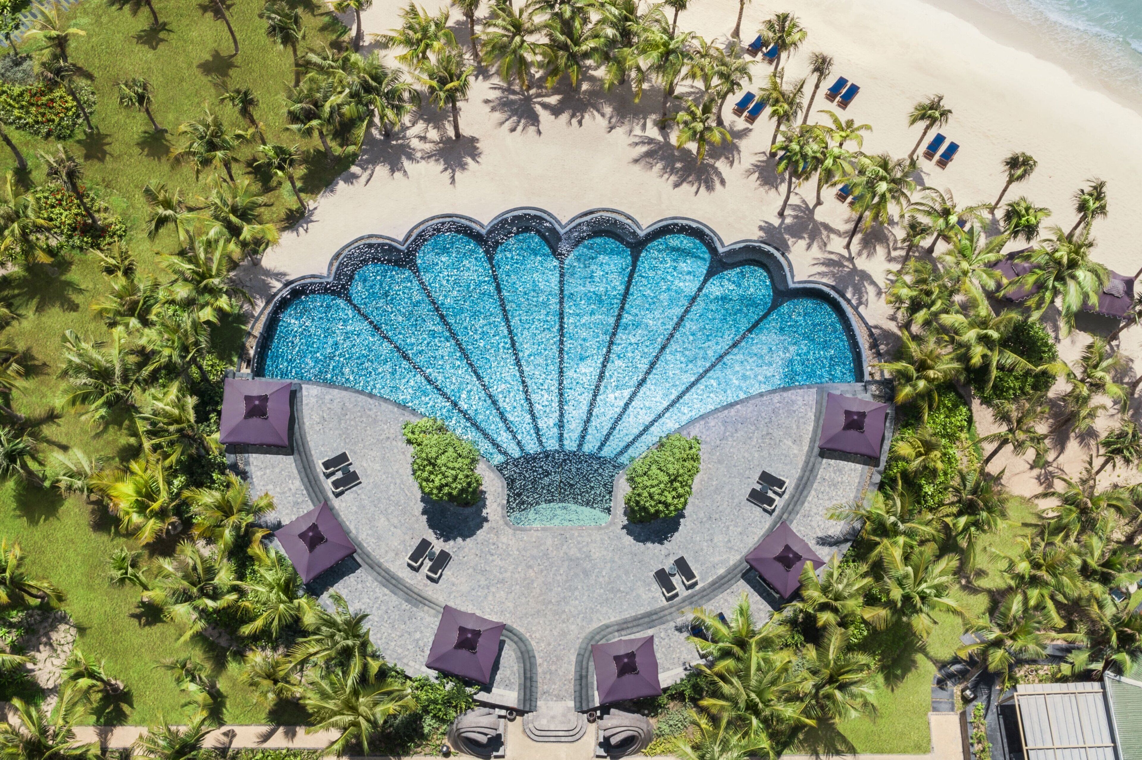 An aerial view of a resort with a large, circular pool surrounded by palm trees and beach chairs.