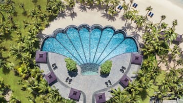 An aerial view of a resort with a large, circular pool surrounded by palm trees and beach chairs.