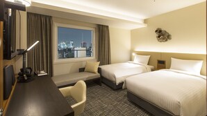 Habitación de hotel con dos camas, un escritorio con silla, televisión y ventana con vistas a la ciudad.