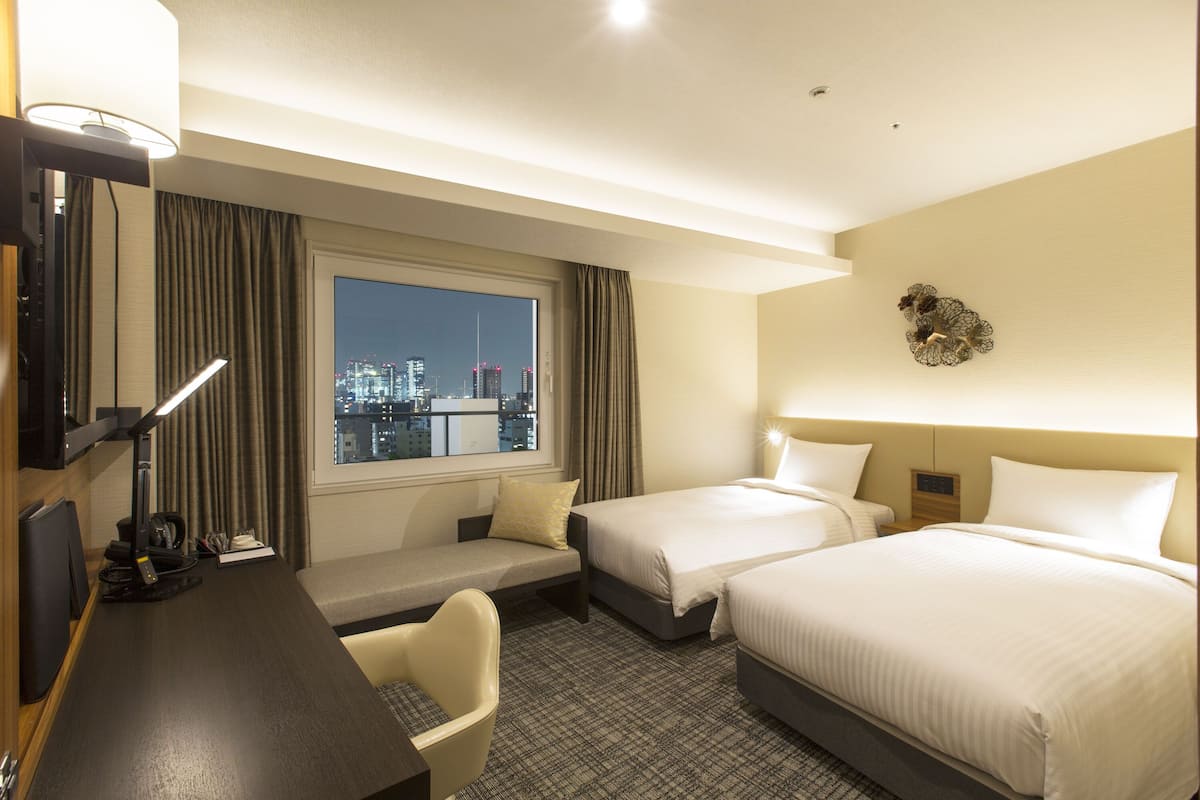 A hotel room with two beds, a desk with a chair, a television, and a window with a city view.