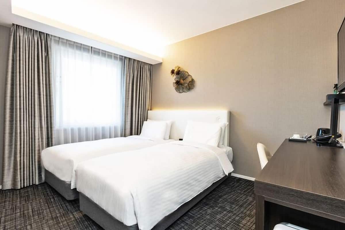 A hotel room with a double bed, a desk with a chair, a large window with curtains, and a wall-mounted TV.