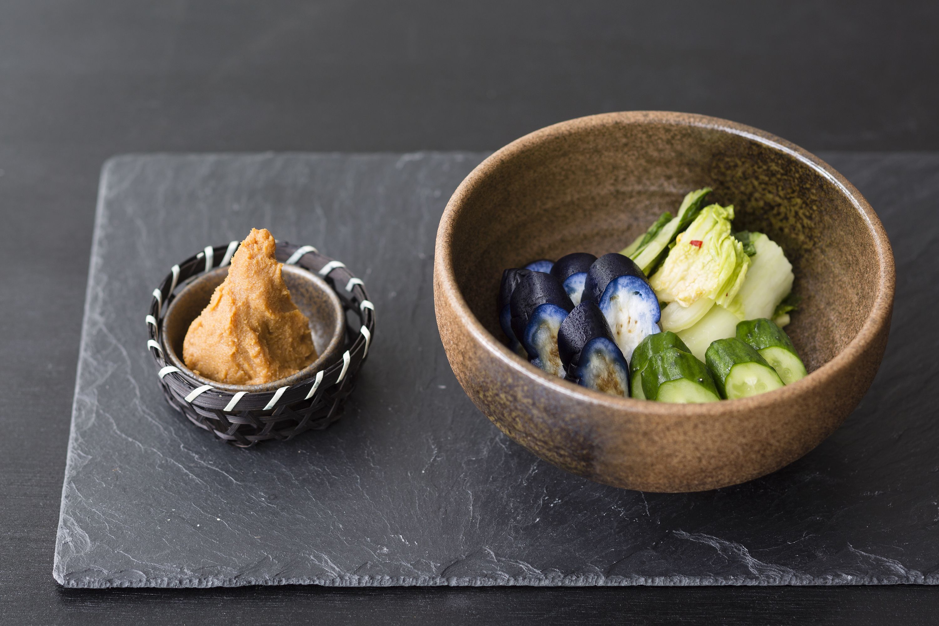 Una tabla de pizarra con un bol de verduras en rodajas y un recipiente pequeño de un condimento marrón.
