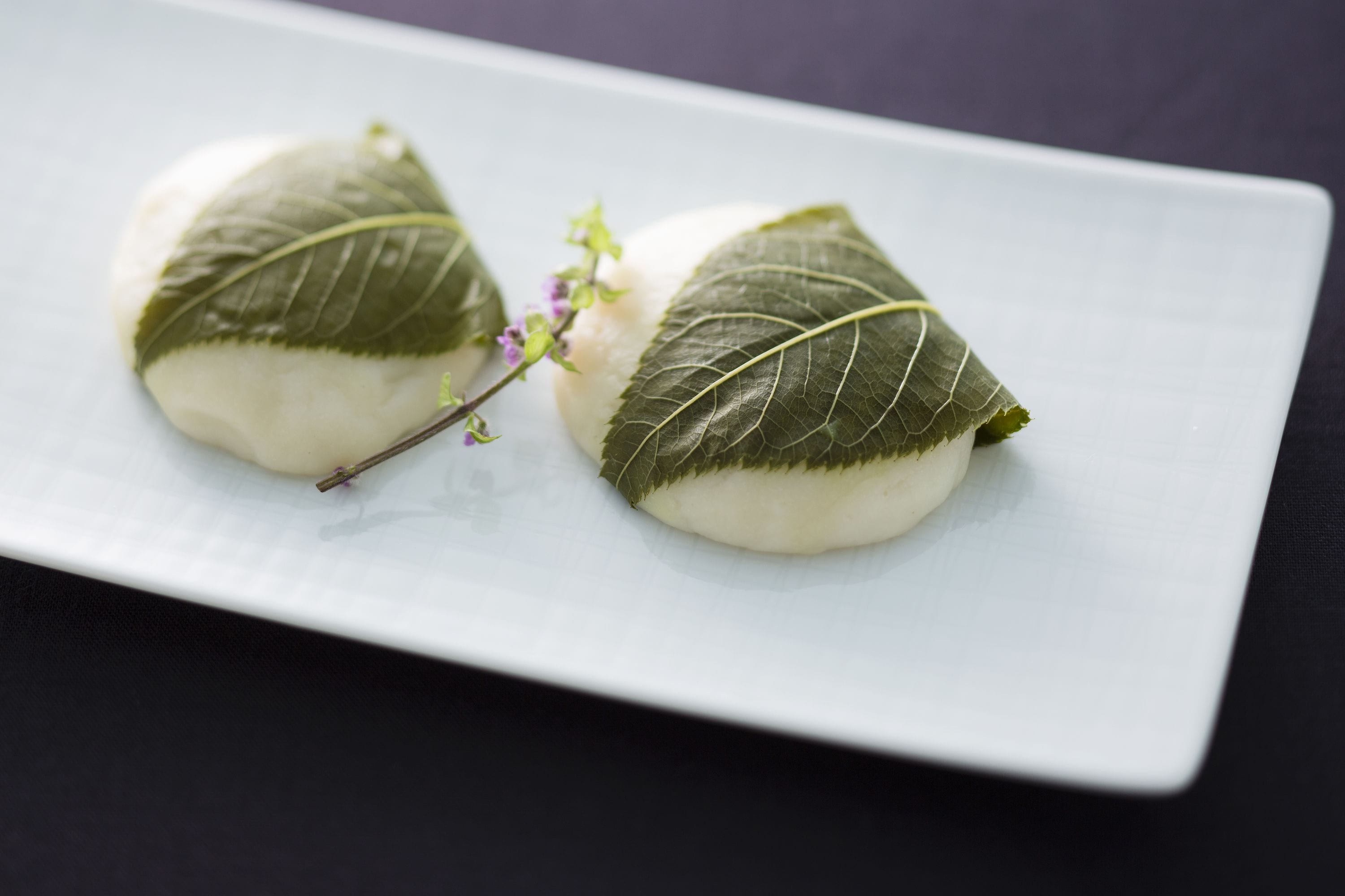 Two dumplings wrapped in green leaves on a white plate.